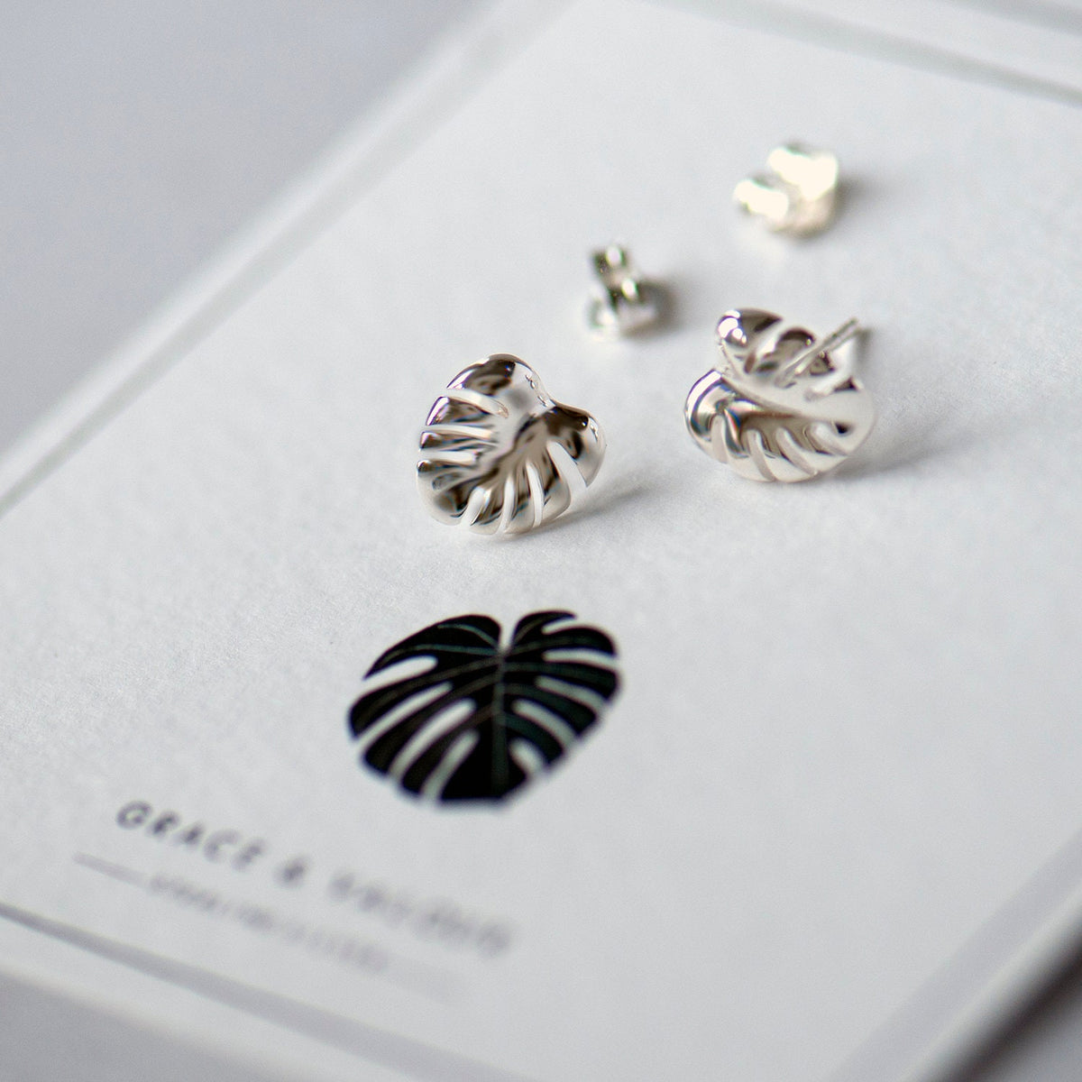 Silver stud earrings designed in the shape of a Monstera leaf, displayed on a white card with the brand name 'Grace & Valour' printed below.