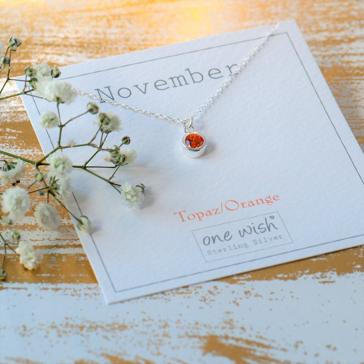 A sterling silver necklace with a small orange topaz heart pendant, displayed on a white card labeled 'November' and 'one wish Sterling Silver', styled with baby's breath flowers on a rustic wooden surface.