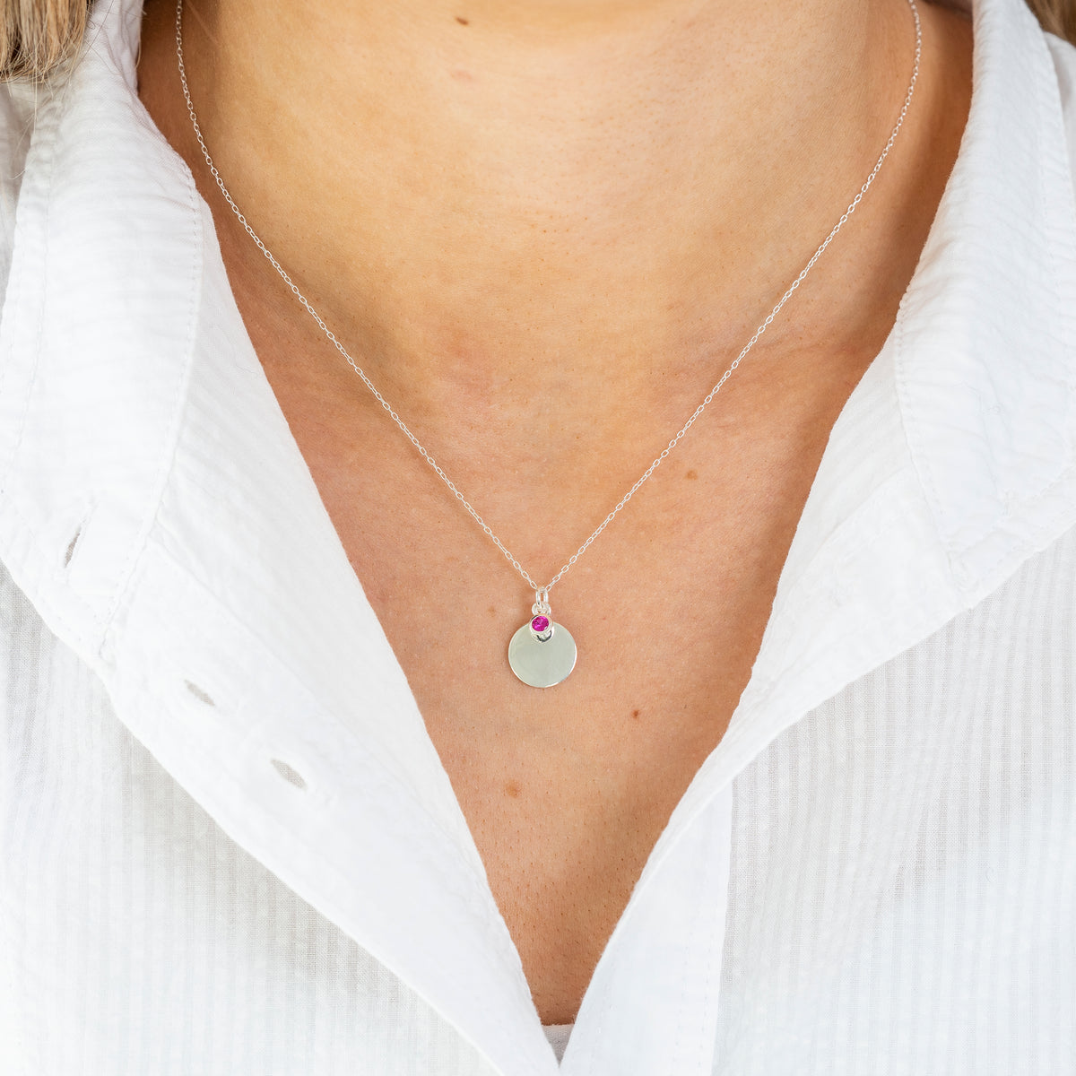 A close-up of a person's neck and chest wearing a delicate silver chain necklace with a small round silver pendant and a tiny pink gemstone accent at the top.