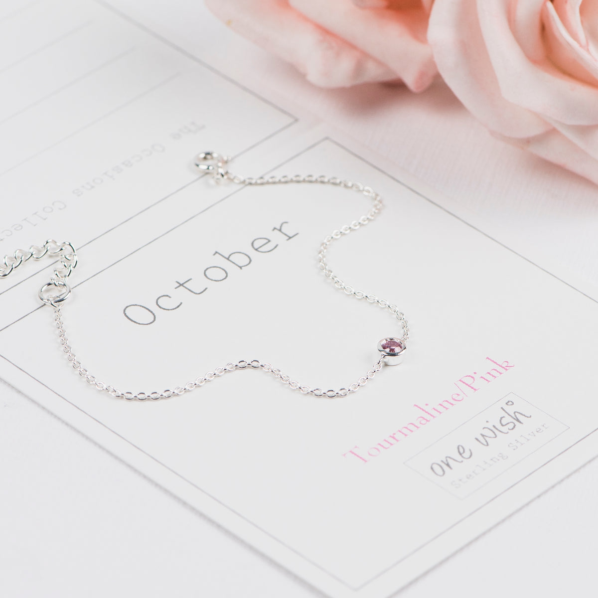 A delicate silver chain bracelet with a small pink gemstone accent, displayed on a white card labeled 'October' with soft pink rose petals in the background.