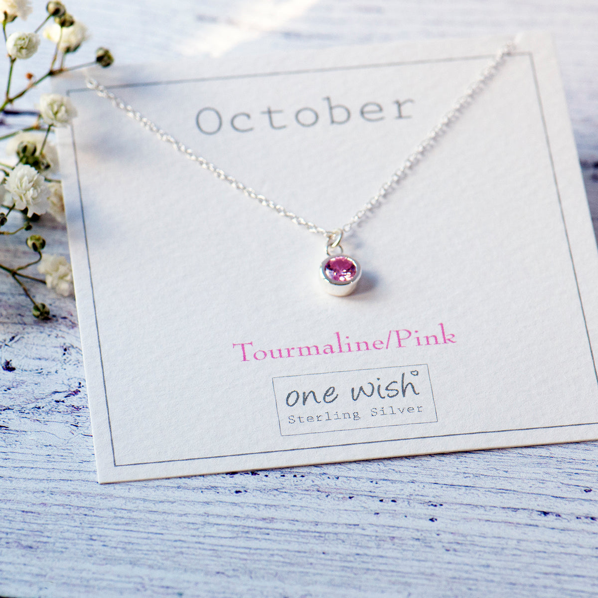 A sterling silver necklace with a pink tourmaline gemstone pendant, displayed on a white card labeled 'October' and 'Tourmaline/Pink' by the brand 'one wish Sterling Silver'. The card is styled on a light blue wooden surface with small white flowers.