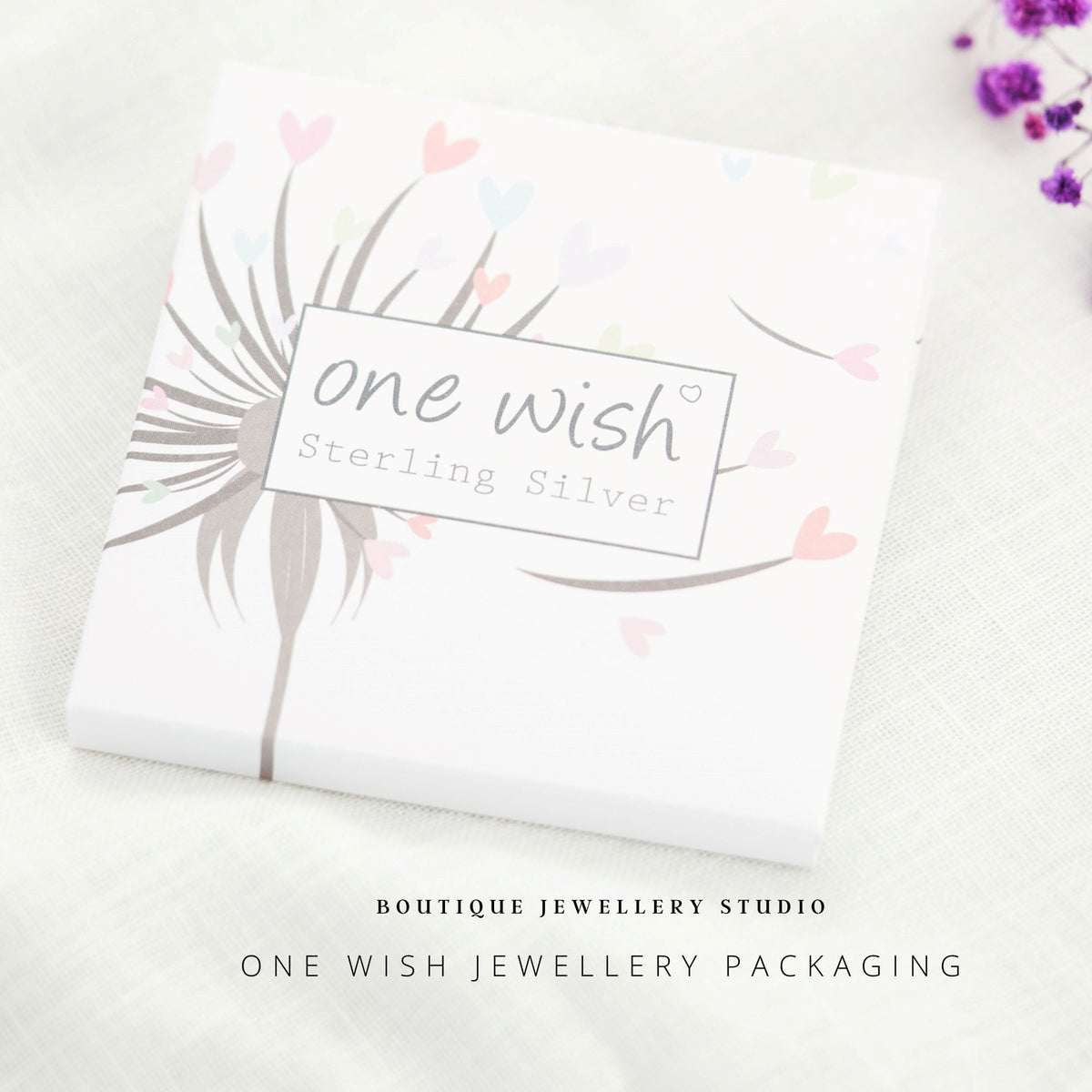 A square white jewelry packaging box with a dandelion illustration and pastel hearts, labeled 'one wish Sterling Silver,' photographed on a light fabric surface with small purple flowers in the corner and boutique text below.
