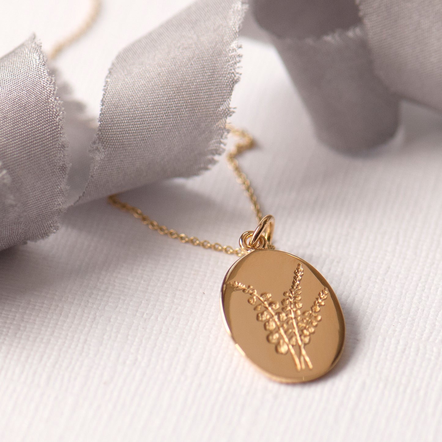 An oval-shaped gold-plated pendant featuring an engraved heather flower design, attached to a gold-plated chain.