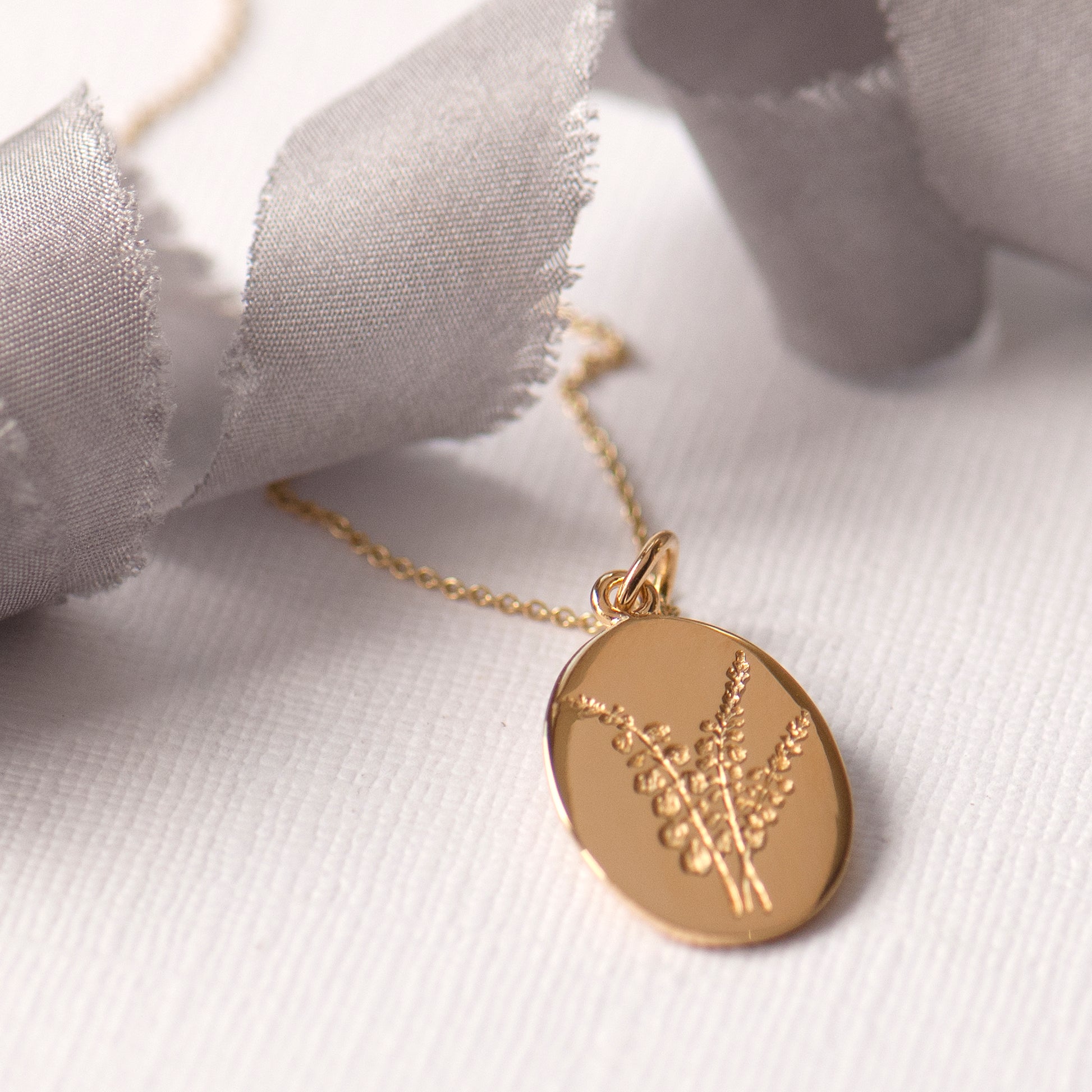 An oval-shaped gold-plated pendant featuring an engraved heather flower design, attached to a gold-plated chain.