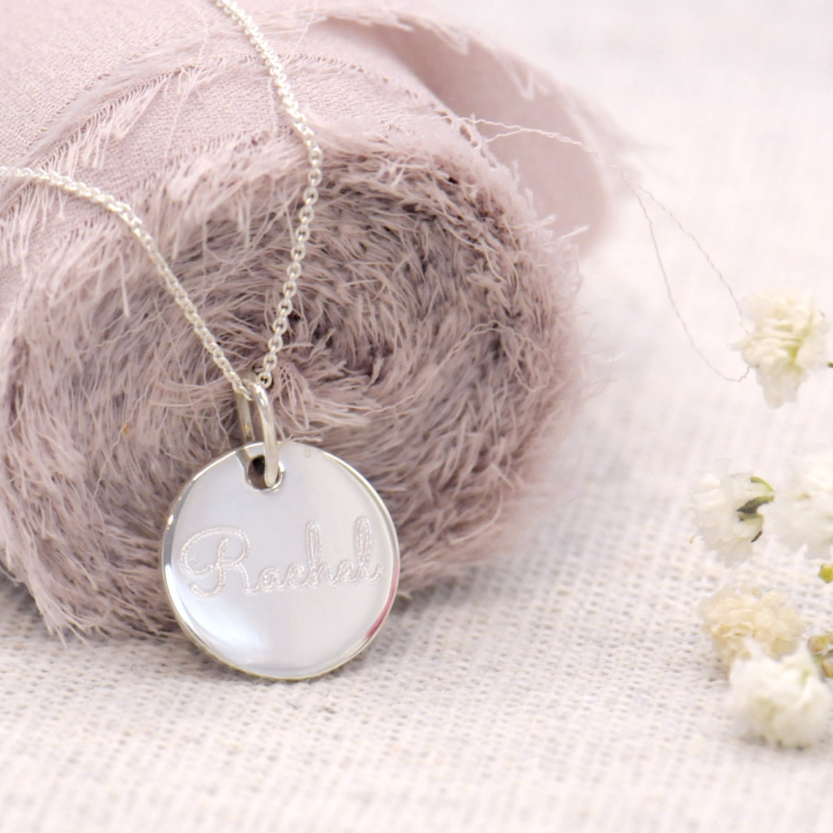 A silver round disc pendant necklace engraved with the name 'Rachel', resting on a dusty mauve fabric roll over a white textured linen surface with small white flowers.