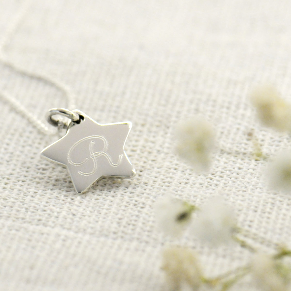 A silver star-shaped pendant necklace with an engraved letter 'R', resting on a textured white fabric background with soft blurred white flowers.