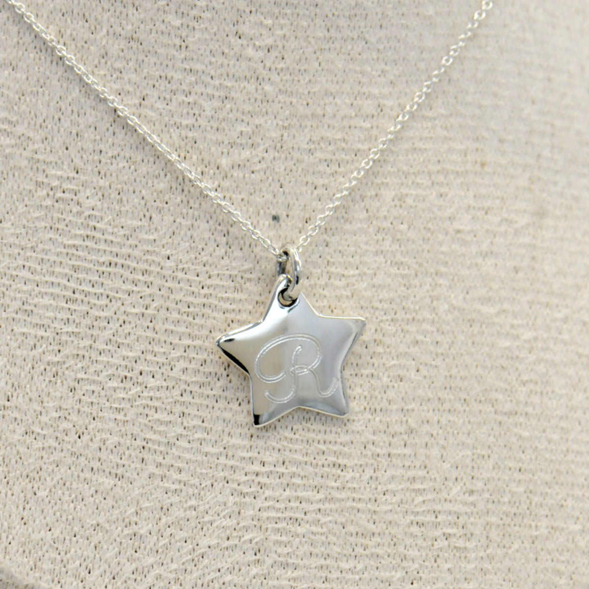 A silver star-shaped pendant necklace with an engraved initial - the letter R, displayed on a textured beige fabric bust.