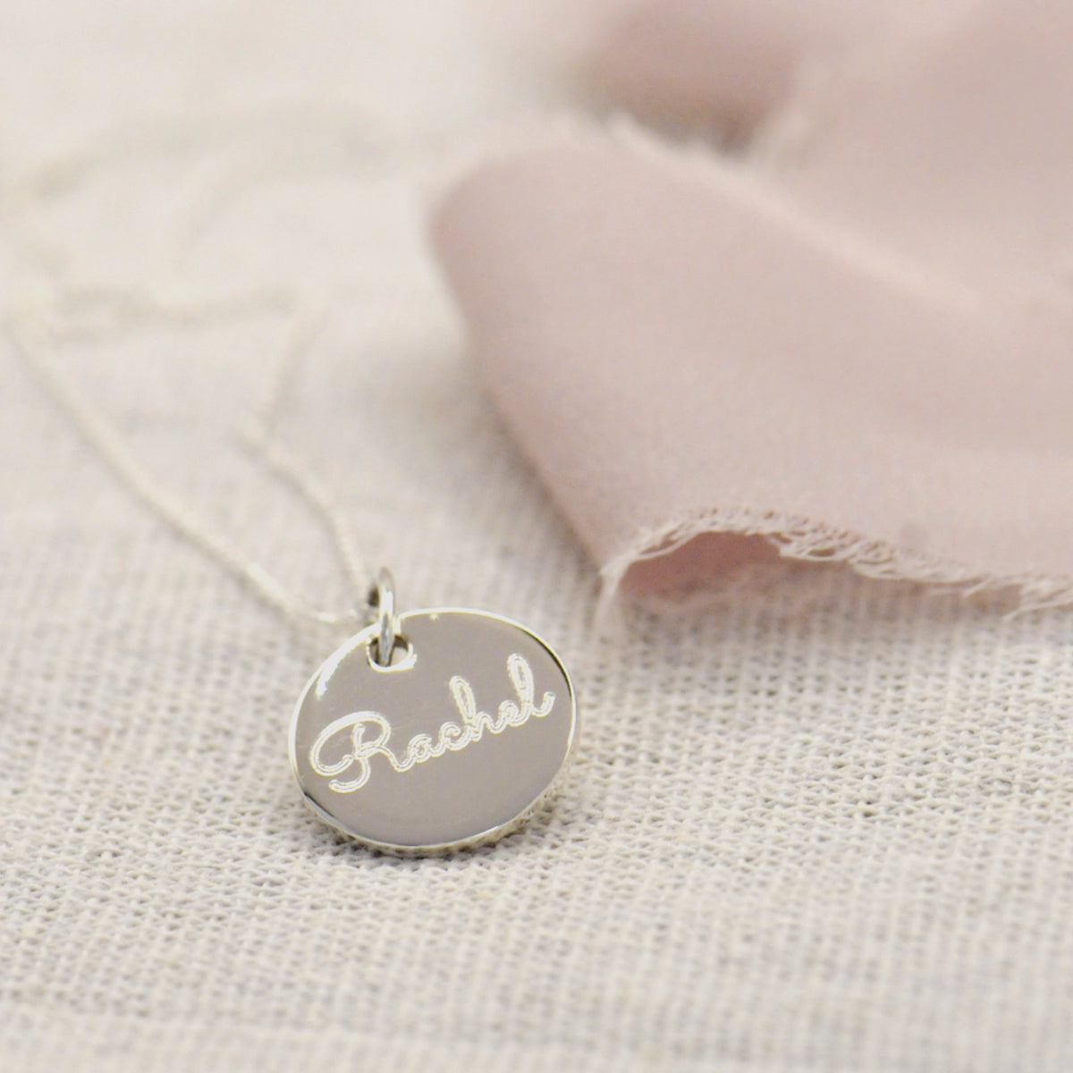 A silver oval pendant necklace engraved with the name 'Rachel' in cursive, resting on a textured beige fabric next to a soft pink cloth.