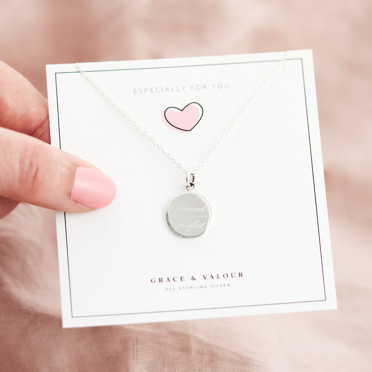 A silver round disc pendant necklace on a delicate chain, displayed on a white gift card held by a hand. The card features a pink heart outline and the text 'GRACE & VALOUR'.