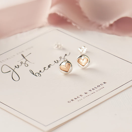 A pair of silver heart stud earrings with gold heart inlays, displayed on a branded jewelry card that reads 'Just because' and 'Grace & Valour'.