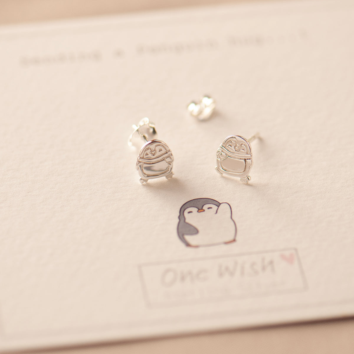 A pair of sterling silver stud earrings designed like penguins, displayed on a white card with a drawing of a penguin and the text 'One Wish'.