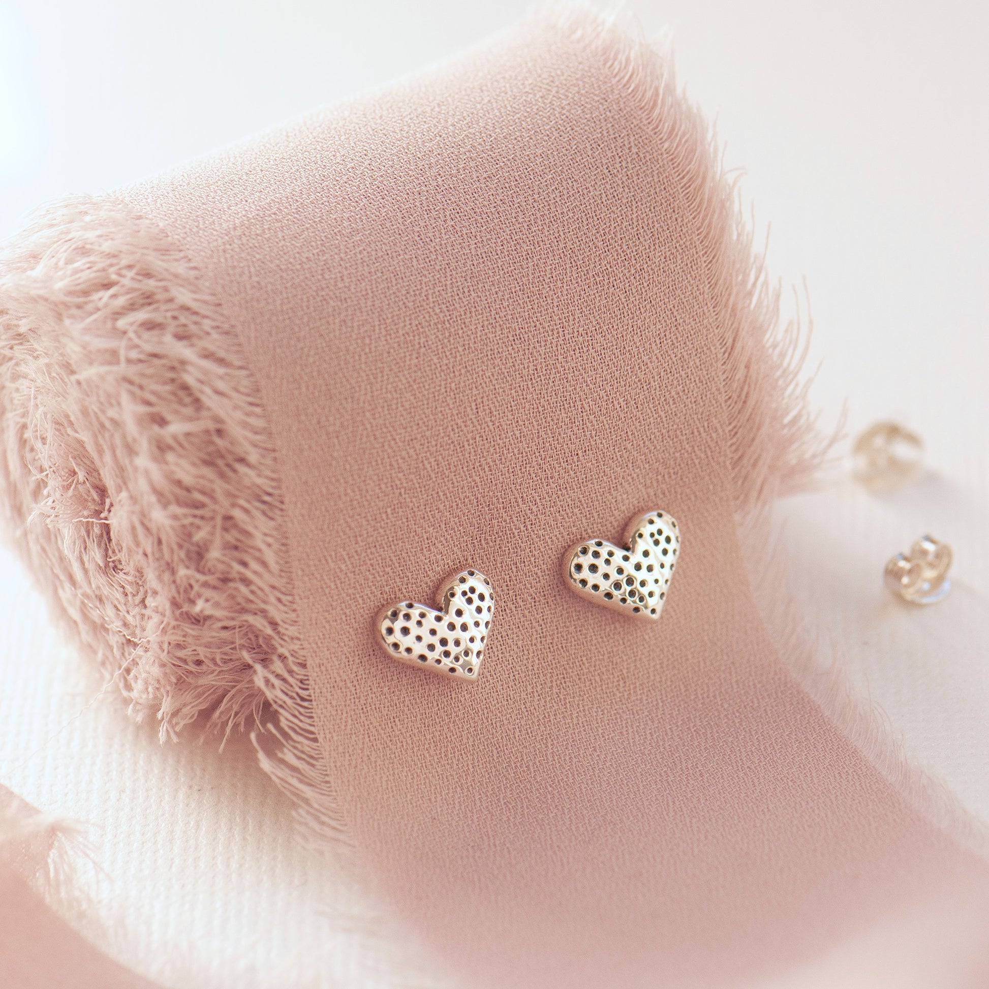 A pair of small heart-shaped stud earrings displayed on a soft blush pink  stand against a light background.