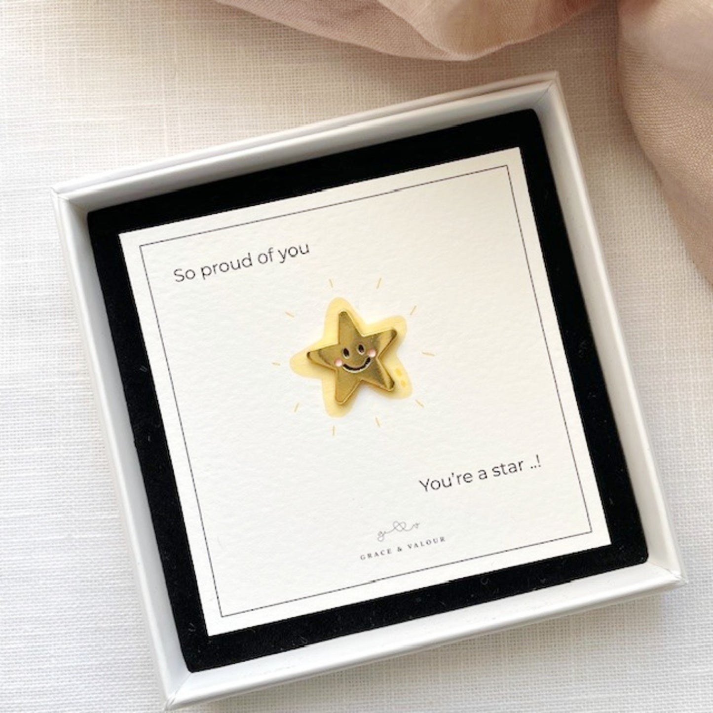 Gold star-shaped pin with a smiley face on a card in a white box, with text 'So proud of you' and 'You're a star'.