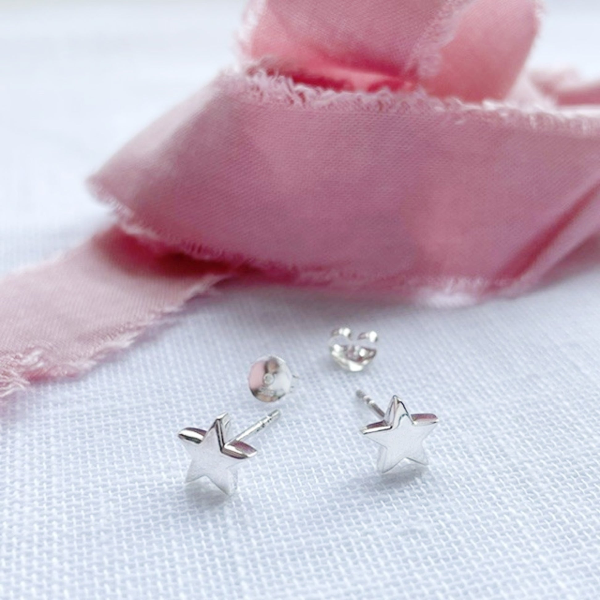 A pair of silver star-shaped stud earrings with butterfly backings, displayed on a textured white fabric next to a soft pink ribbon.