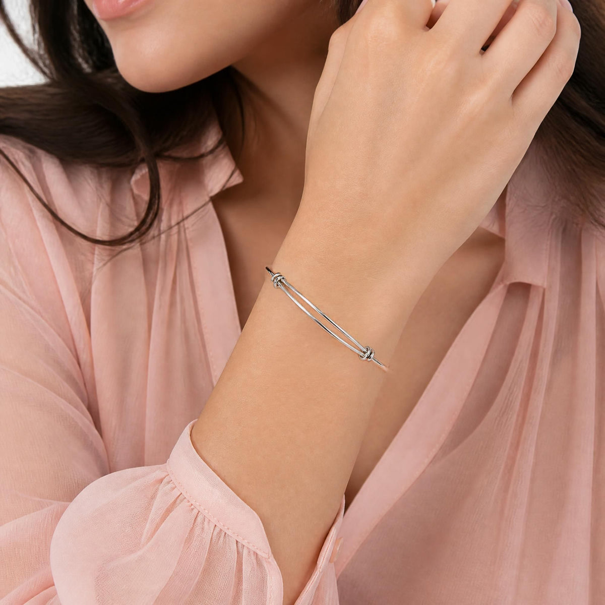 A close-up of a woman wearing a delicate silver-toned double-bar bangle bracelet with small bead details at the ends, paired with a soft pink blouse.
