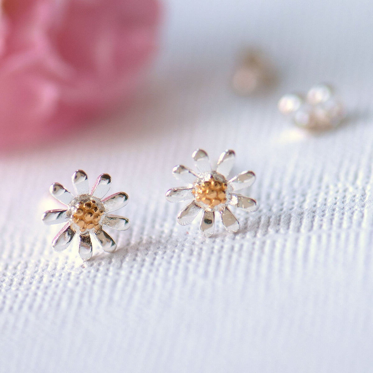 A pair of sterling silver daisy stud earrings with gold centers, displayed on a white textured surface with a pink flower in the background.