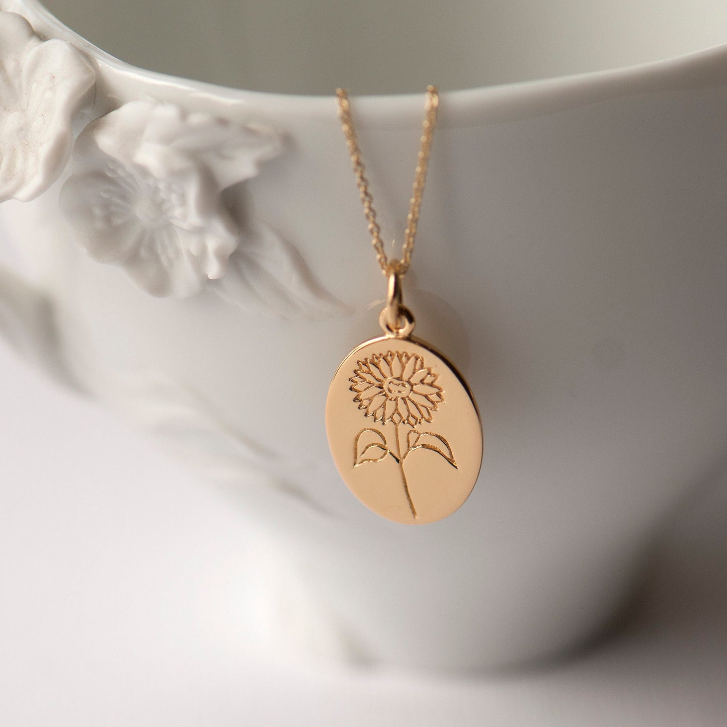 A gold-plated sunflower pendant necklace with an oval disc-shaped pendant, displayed on a white floral-patterned stand.