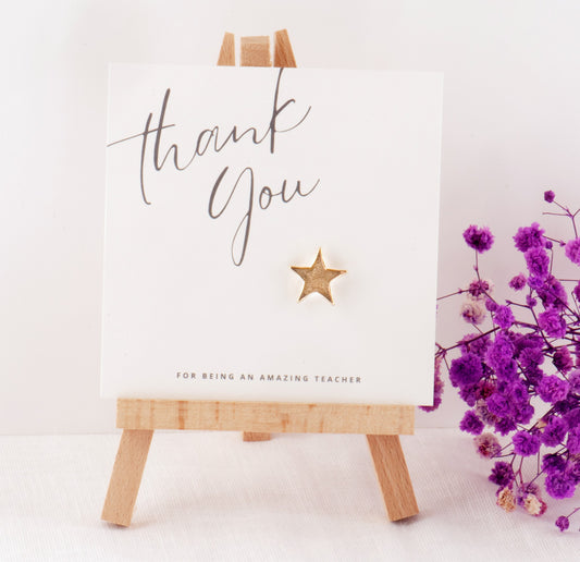 White card with 'thank you for being an amazing teacher' text on a wooden easel with purple flowers.