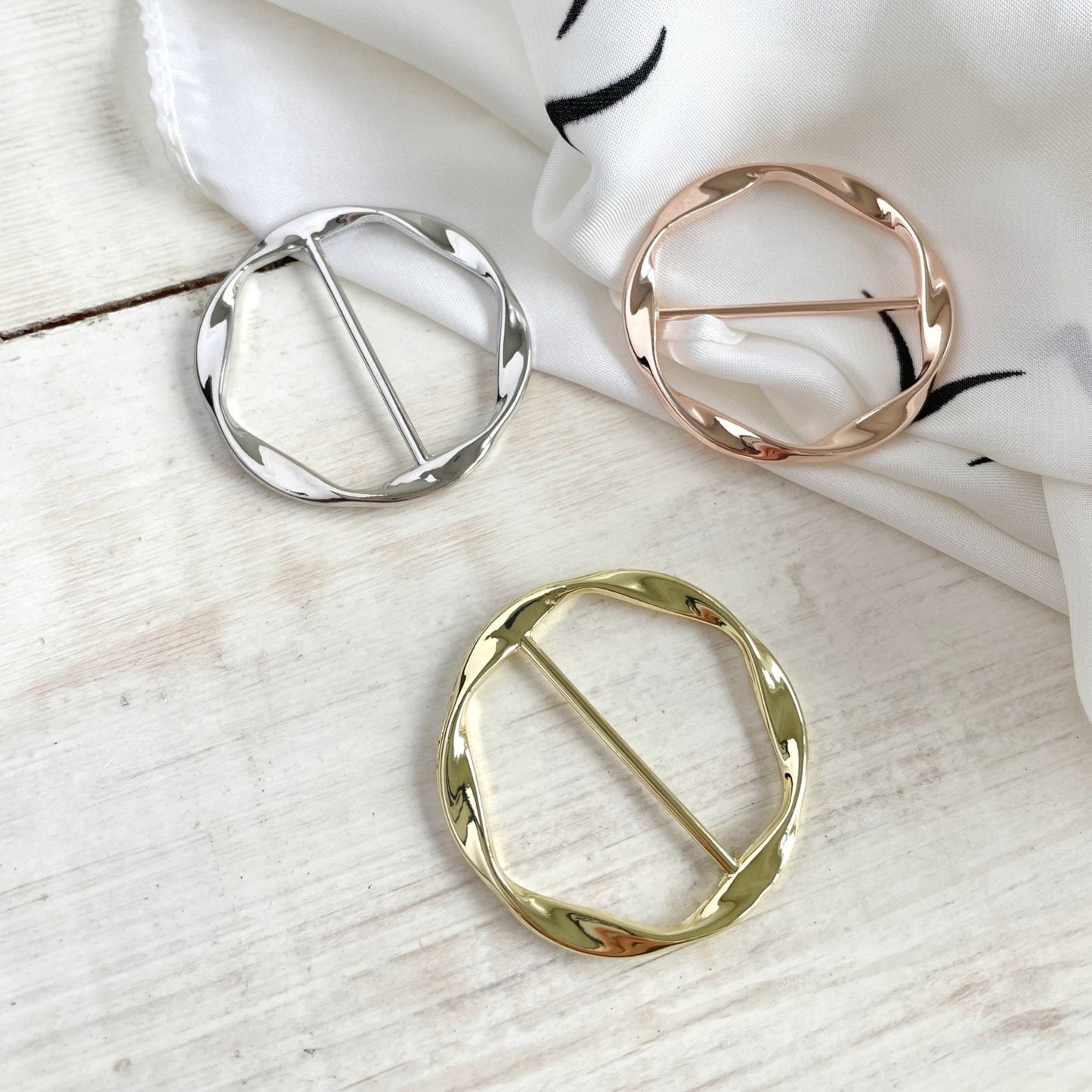 Three metallic scarf rings in gold, silver, and rose gold on a white fabric background.