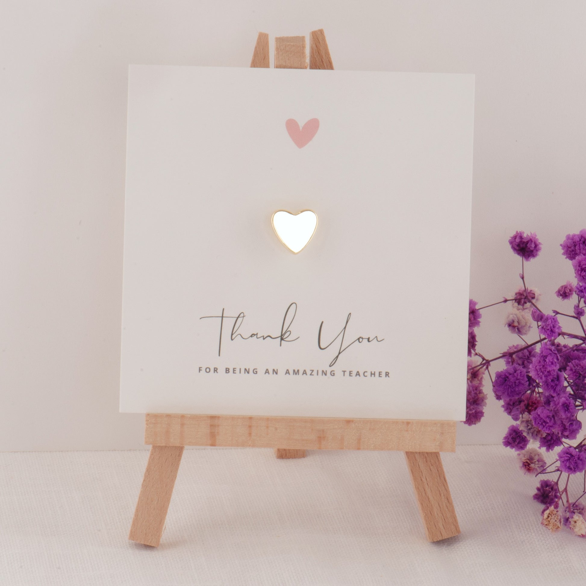 Card on a wooden easel with 'Thank you for being an amazing teacher' text and heart designs, next to purple flowers.