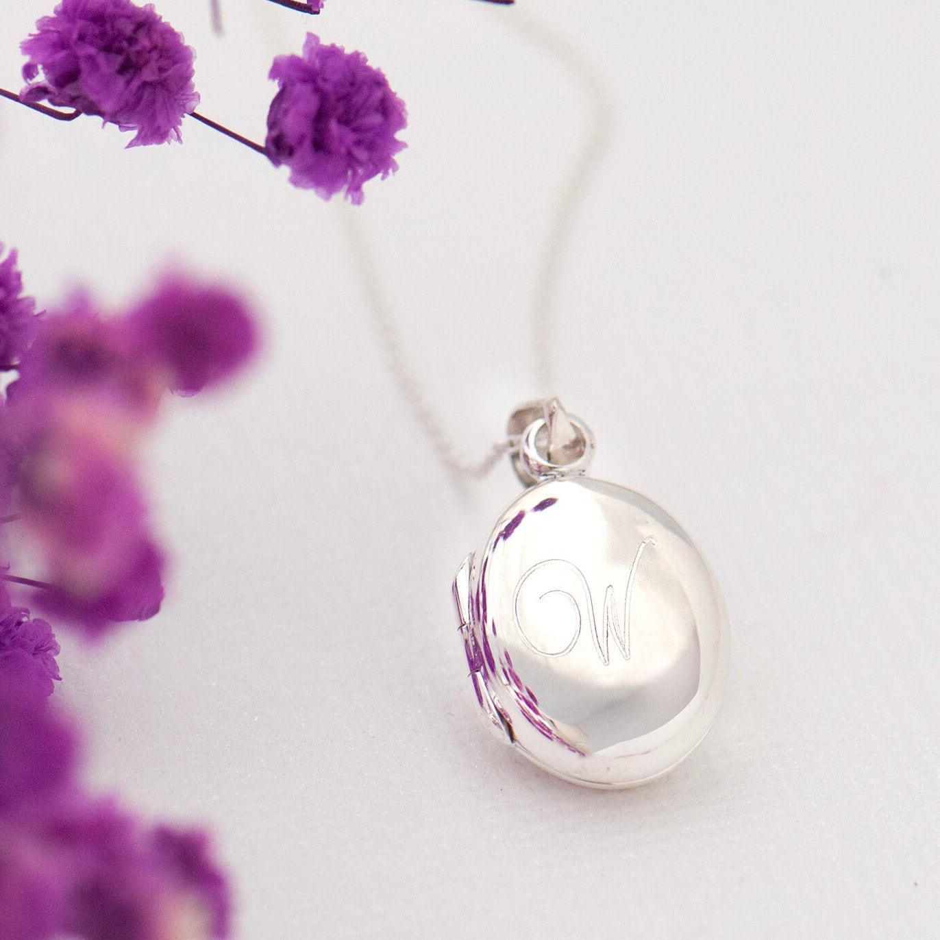 A high-polished sterling silver oval locket engraved with an initial necklace with a chain, displayed against a background of purple flowers.