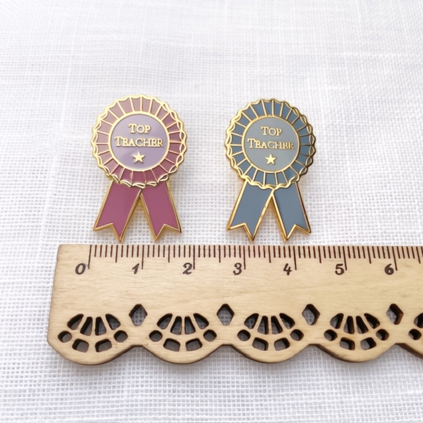 Two 'Top Teacher' pins with ribbons on a white background, one pink and one blue, next to a wooden ruler.
