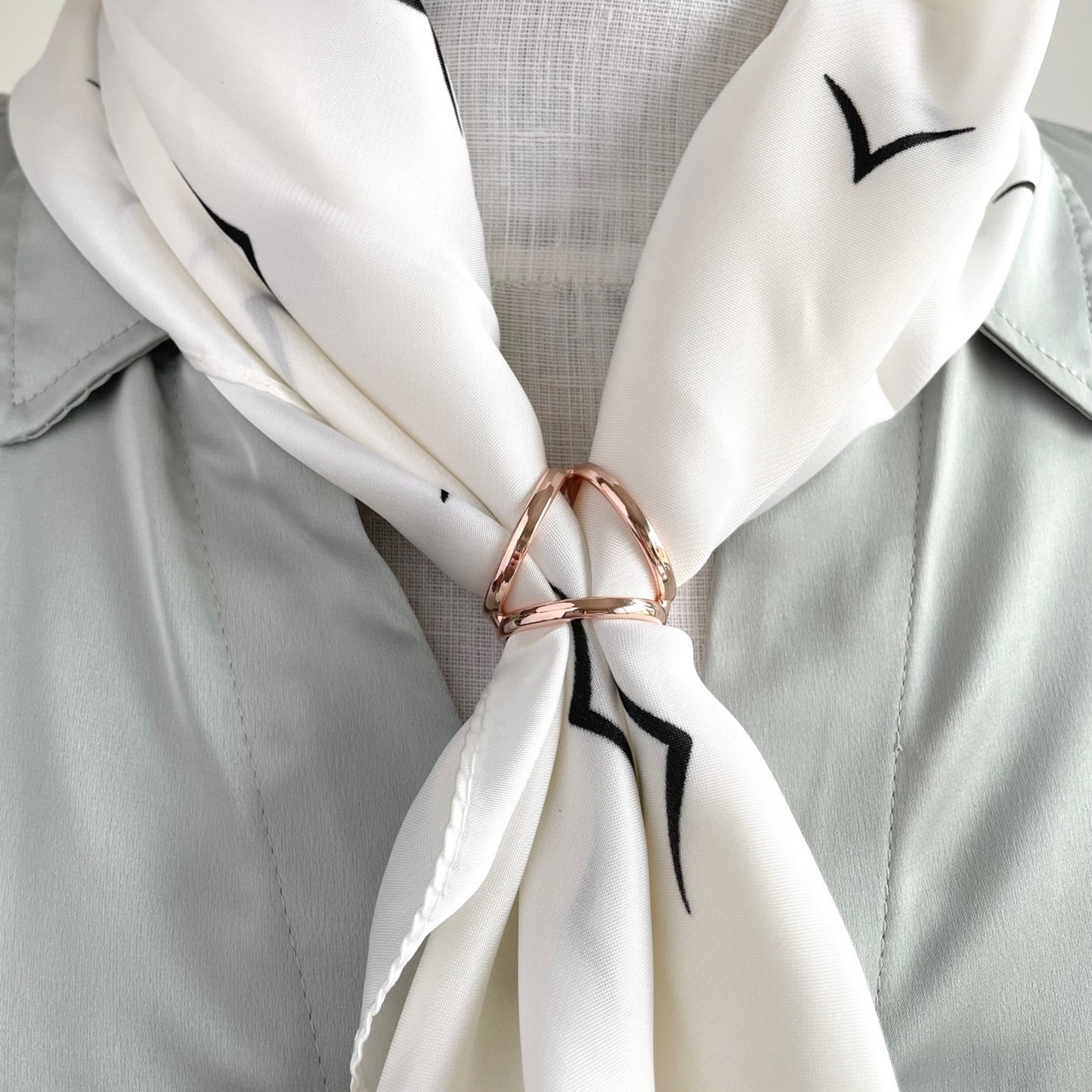 White scarf with black bird pattern tied around a gray shirt collar, held by a rose gold ring.