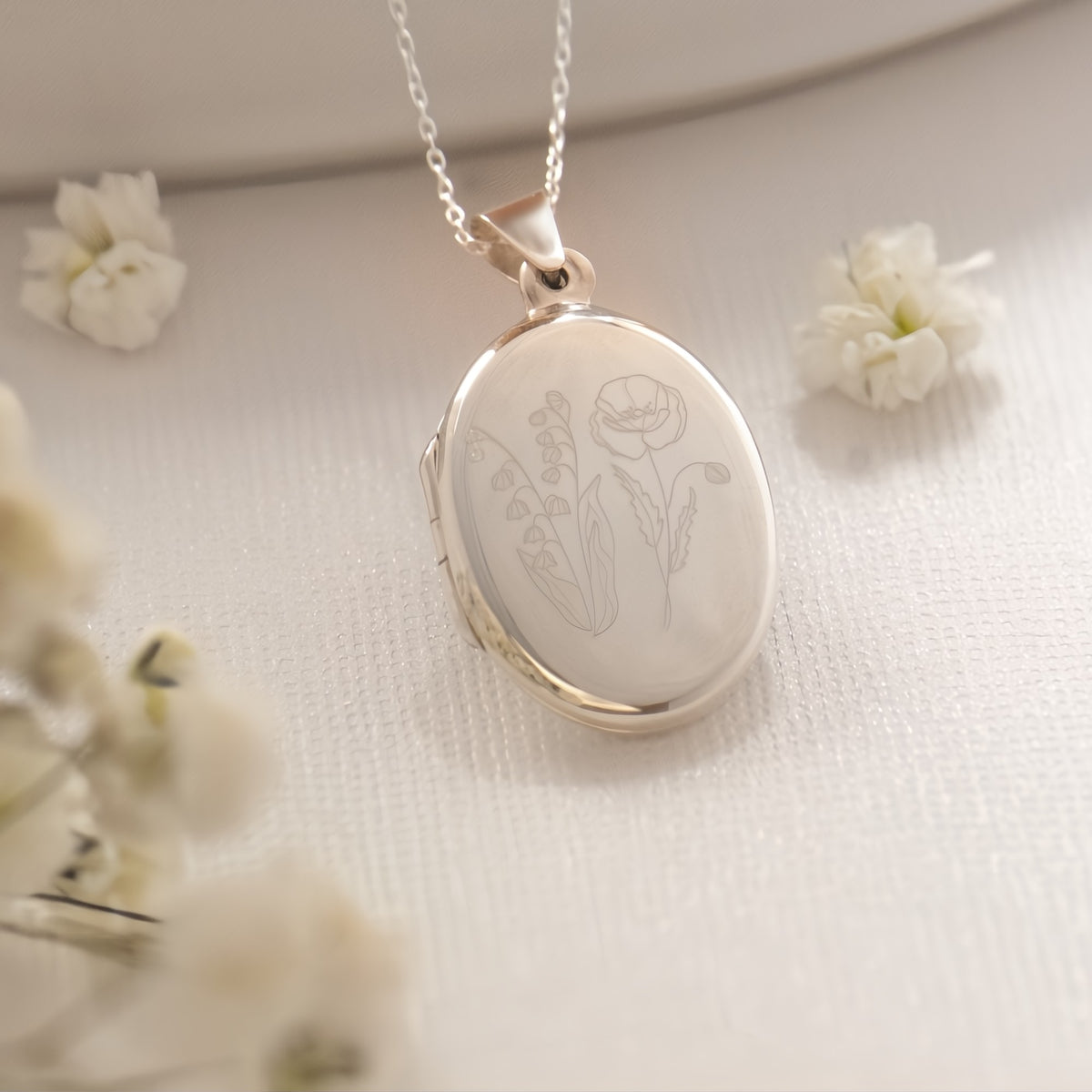 An oval locket pendant necklace with a silver-toned chain, featuring an engraved floral design of a Poppy and lily of the valley. The pendant is displayed on a textured cream fabric background with soft white flowers in the foreground and background.