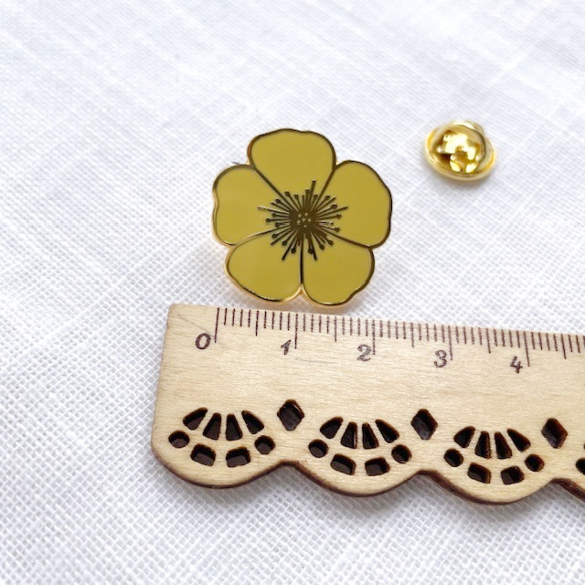 A small flower-shaped gold pin with a yellow center is shown next to a wooden ruler for scale on a white textured fabric background.