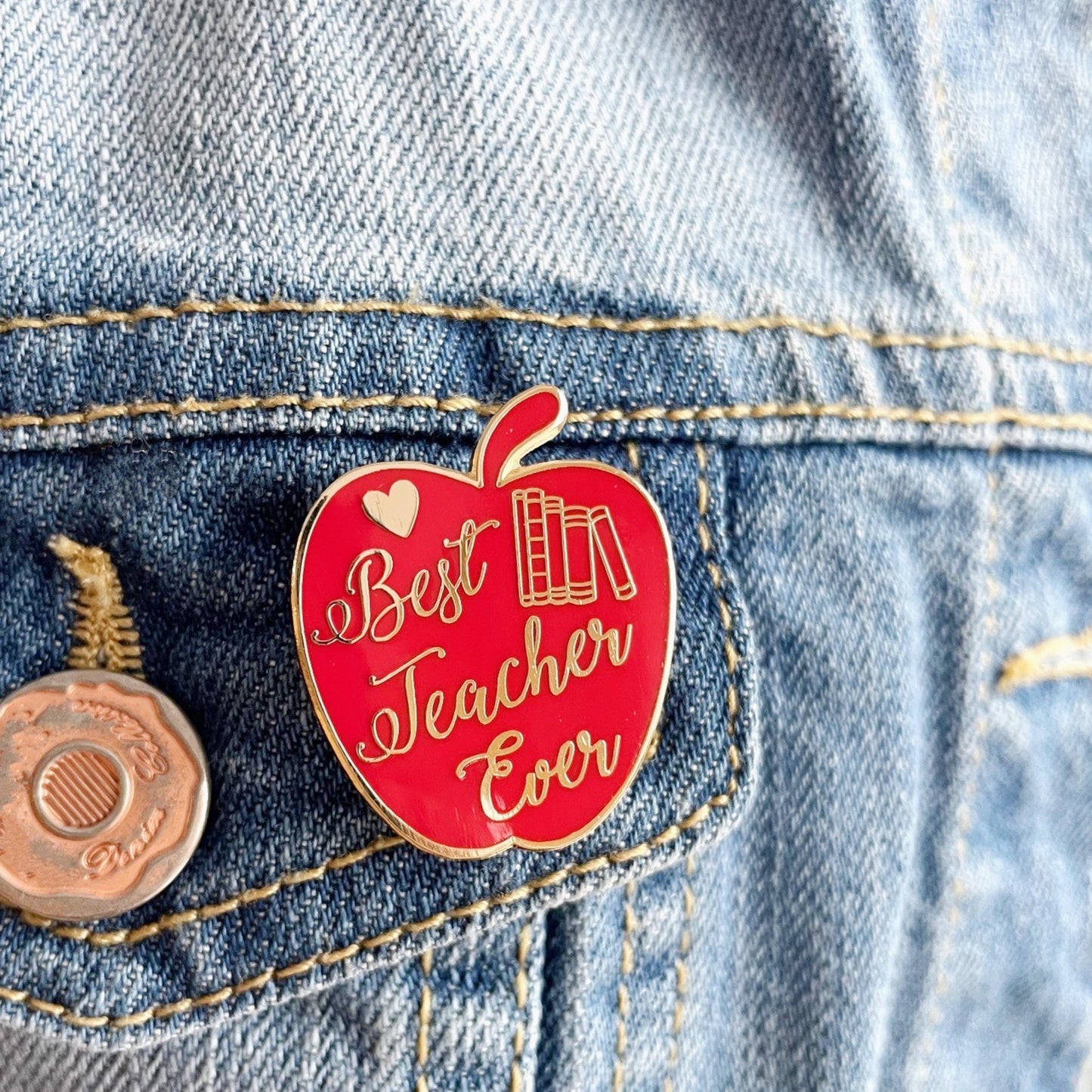 Red apple-shaped pin with 'Best Teacher Ever' text on a denim jacket.