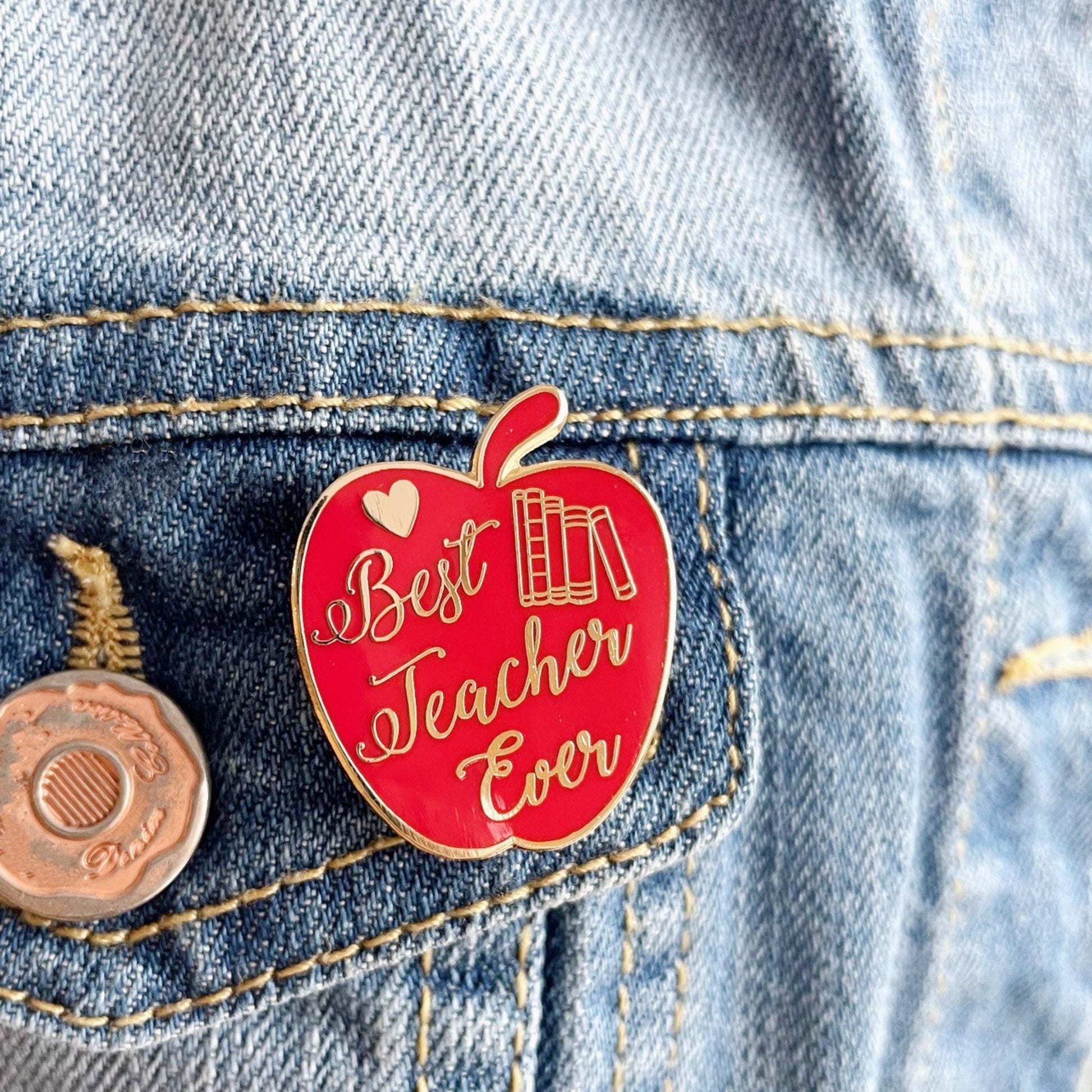Red apple-shaped pin with 'Best Teacher Ever' text on a denim jacket.