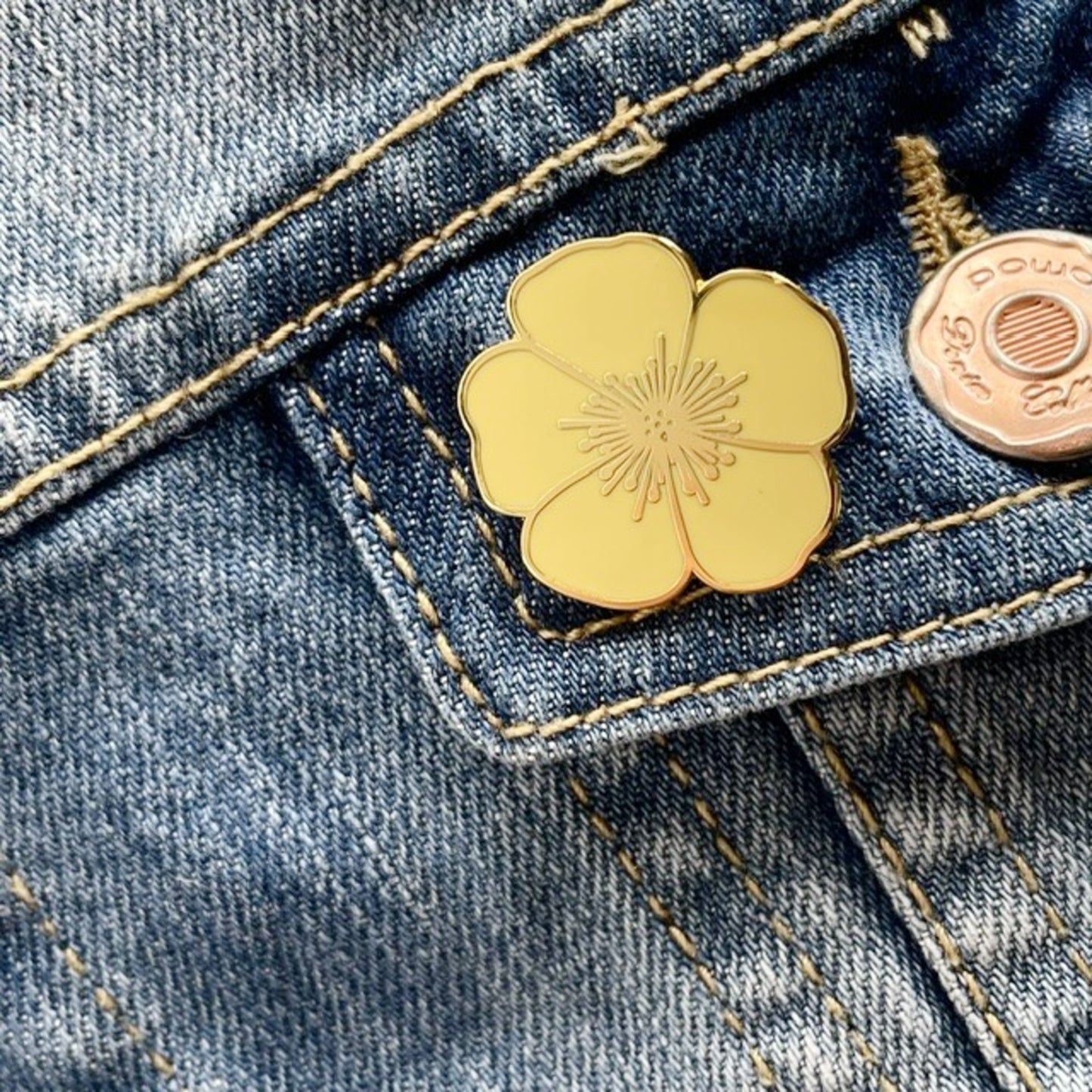 A close-up of a yellow-tone flower-shaped enamel pin attached to a blue denim pocket.