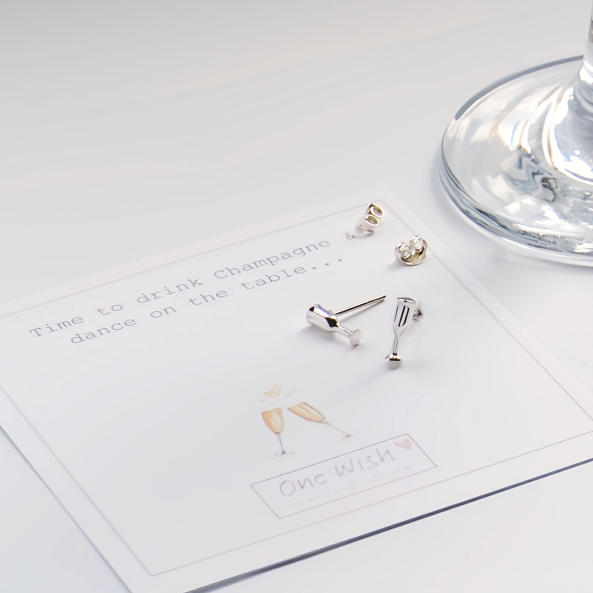 A pair of silver-toned stud earrings shaped like champagne glasses and a small champagne bottle, displayed on a printed card that reads 'Time to drink Champagne dance on the table.