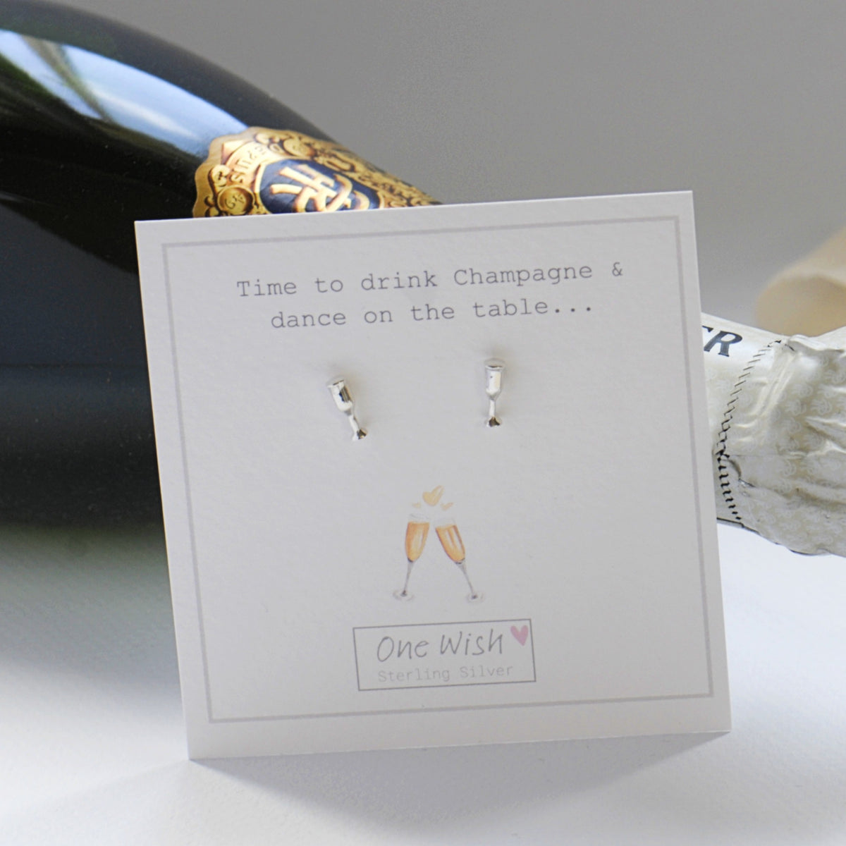 A pair of small silver stud earrings displayed on a white card that reads 'Time to drink Champagne & dance on the table.