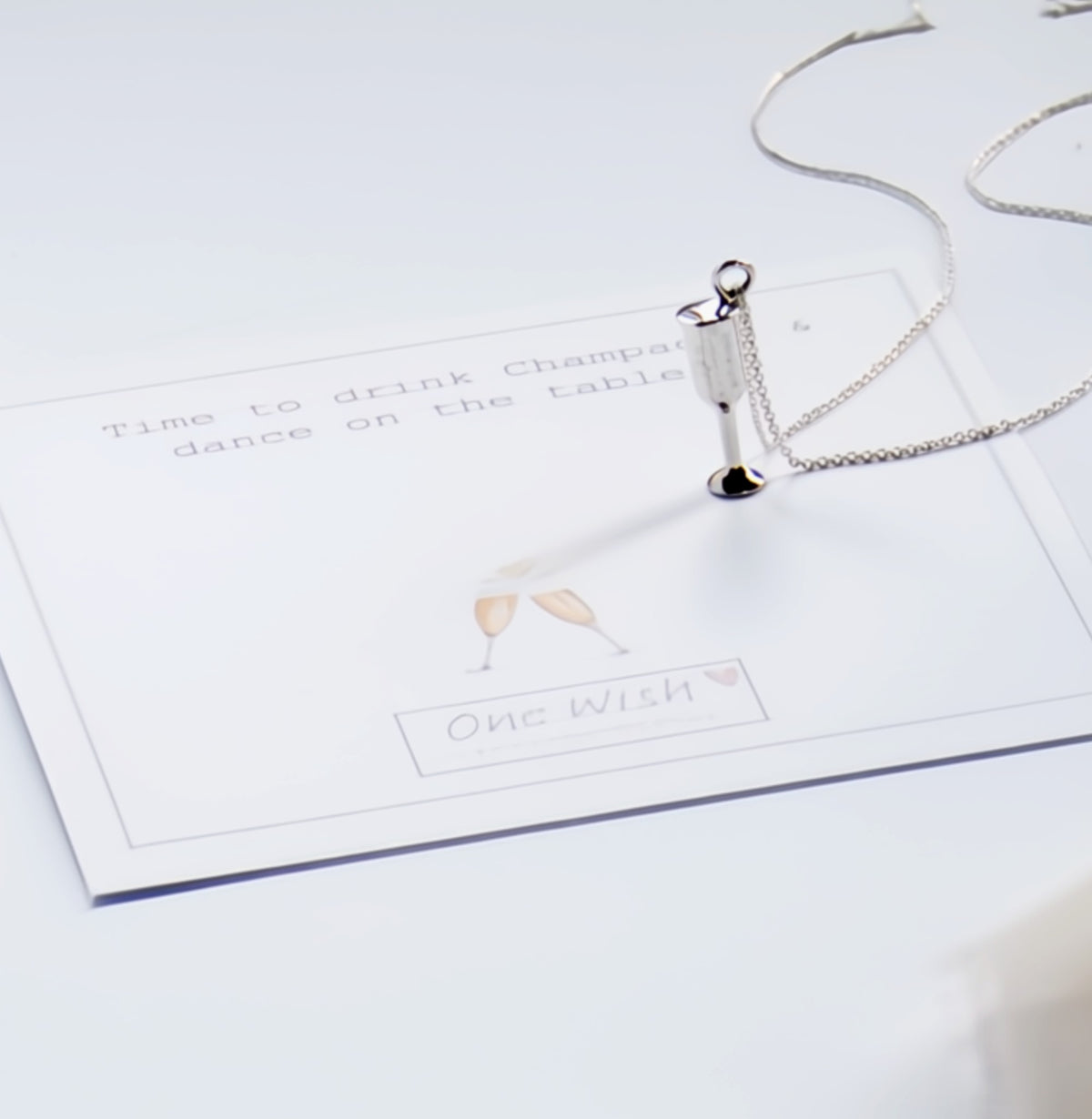 A silver-toned necklace with a small charm shaped like a champagne bottle and clinking glasses, resting on a white printed card that reads 'Time to drink Champagner dance on the Table' and 'One Wish'.