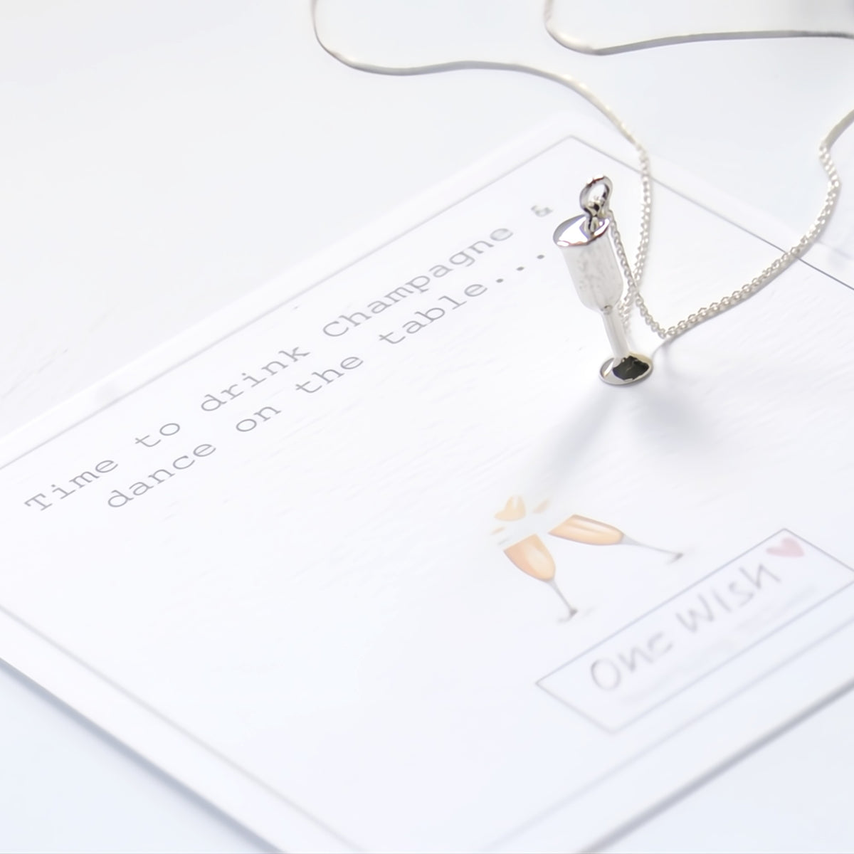 A silver pendant necklace shaped like a champagne bottle lies on a white card with printed text and a small champagne illustration. The necklace is photographed in a clean, bright studio setting.