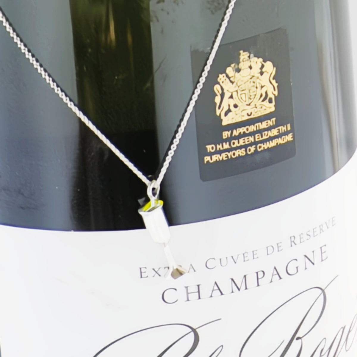 A silver necklace with a small champagne bottle pendant is draped over a bottle of champagne. The champagne bottle has a black label with a gold crest and text reading 'BY APPOINTMENT TO HM QUEEN ELIZABETH II PURVEYORS OF CHAMPAGNE'.