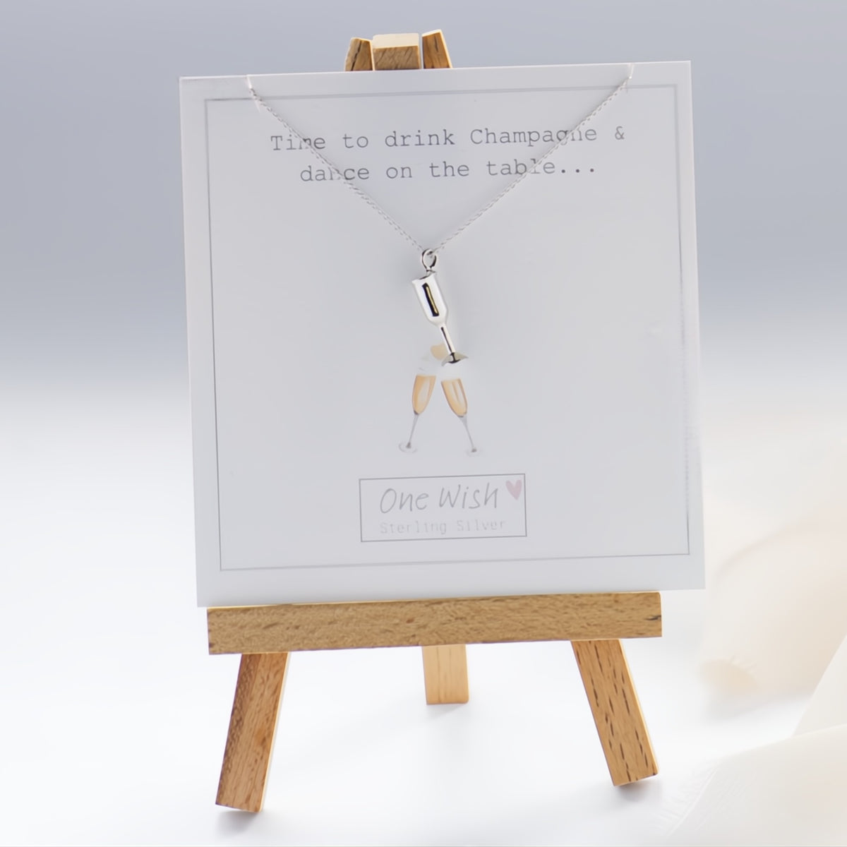 A silver necklace with a small bottle-shaped pendant displayed on a white card mounted on a small wooden easel. The card reads 'Time to drink Champagne & dance on the table.