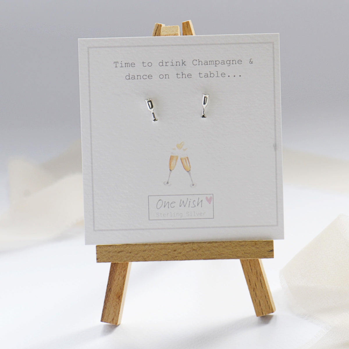 A pair of small silver stud earrings displayed on a white card mounted on a small wooden easel. The card has printed text reading 'Time to drink Champagne & dance on the table.