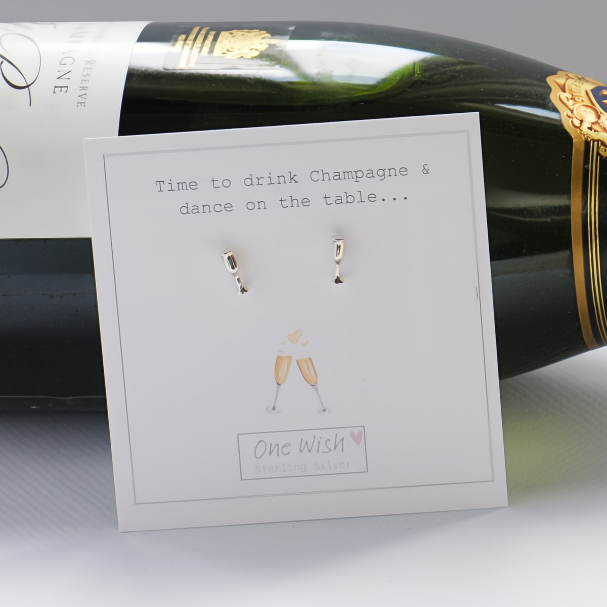 A pair of small silver champagne glass earrings displayed on a white card that reads 'Time to drink Champagne & dance on the table.