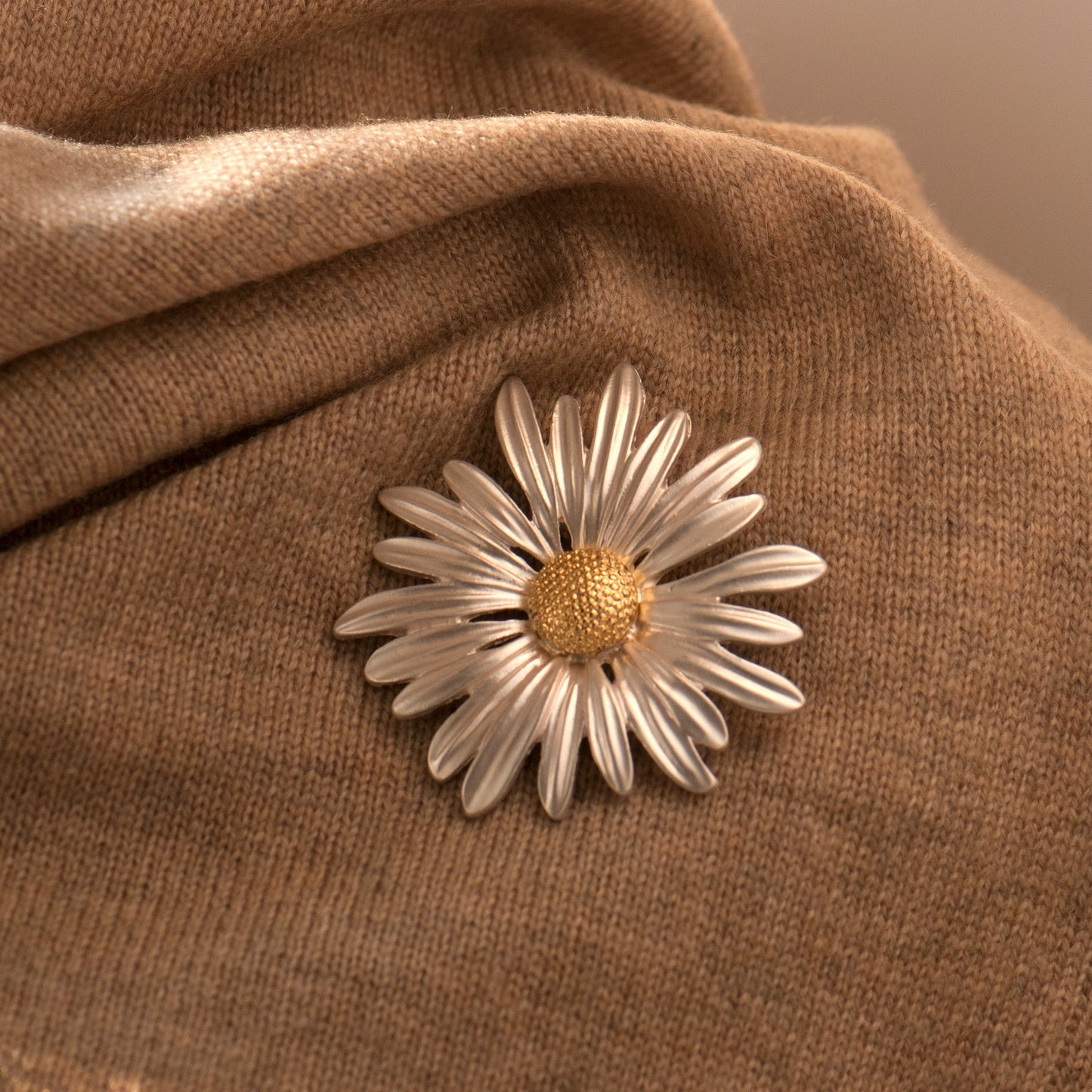 A daisy brooch with silver arching petals and a gold center pinning onto a brown fabric surface.