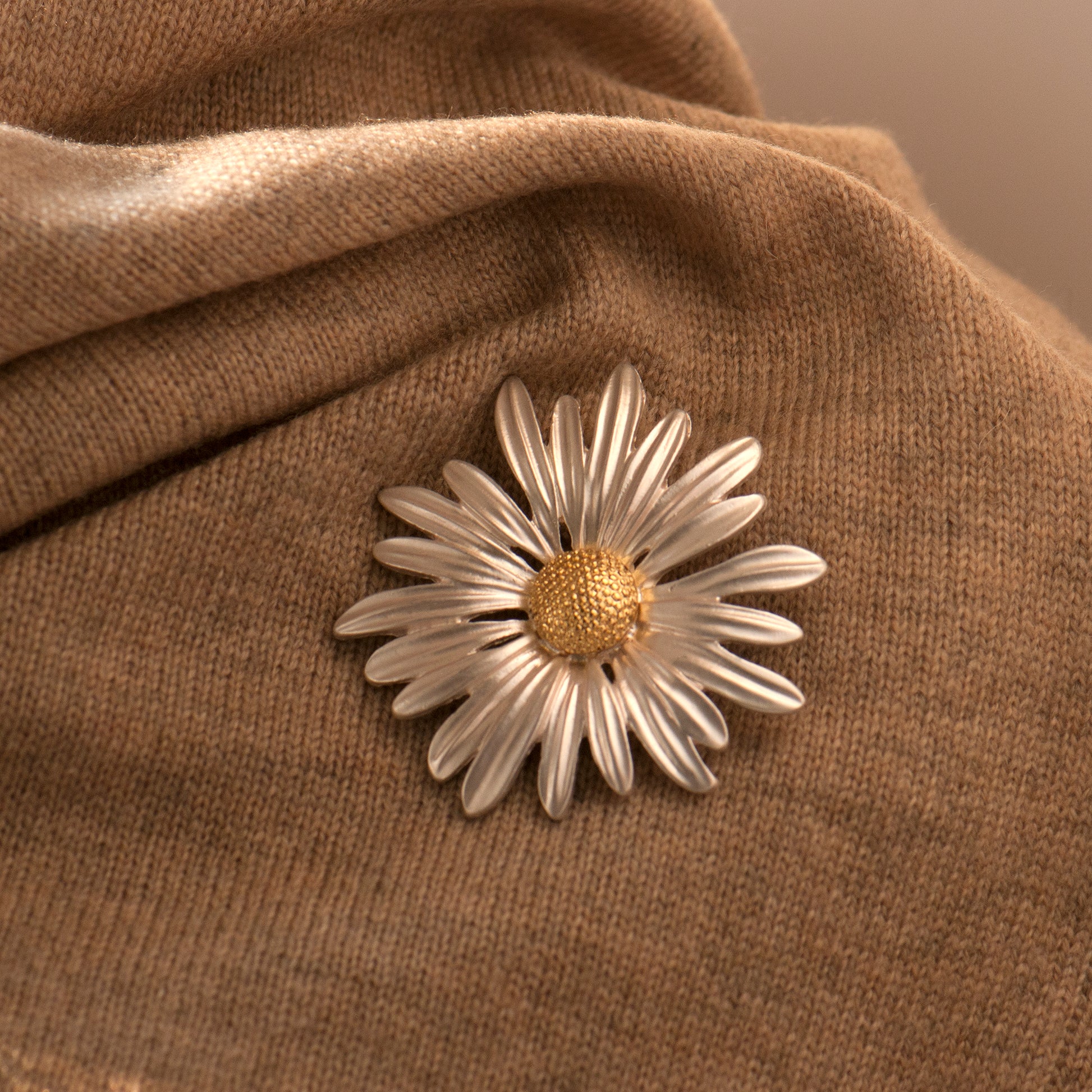 A daisy brooch with silver arching petals and a gold center pinning onto a brown fabric surface.