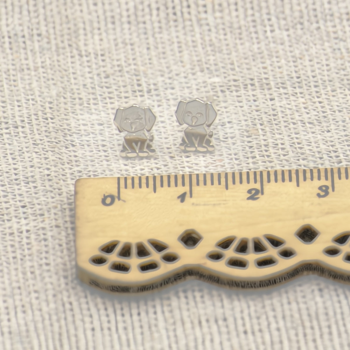 Two small silver stud earrings shaped like puppy dogs, displayed on a textured white fabric next to a wooden ruler for scale.