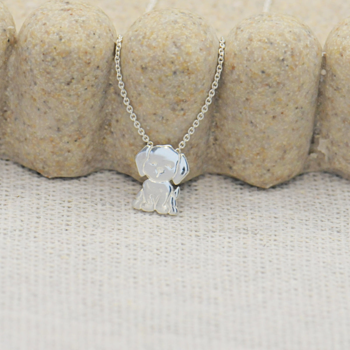 A silver pendant necklace featuring a small, polished dog silhouette charm on a delicate silver chain, displayed against a textured beige fabric and stone background.