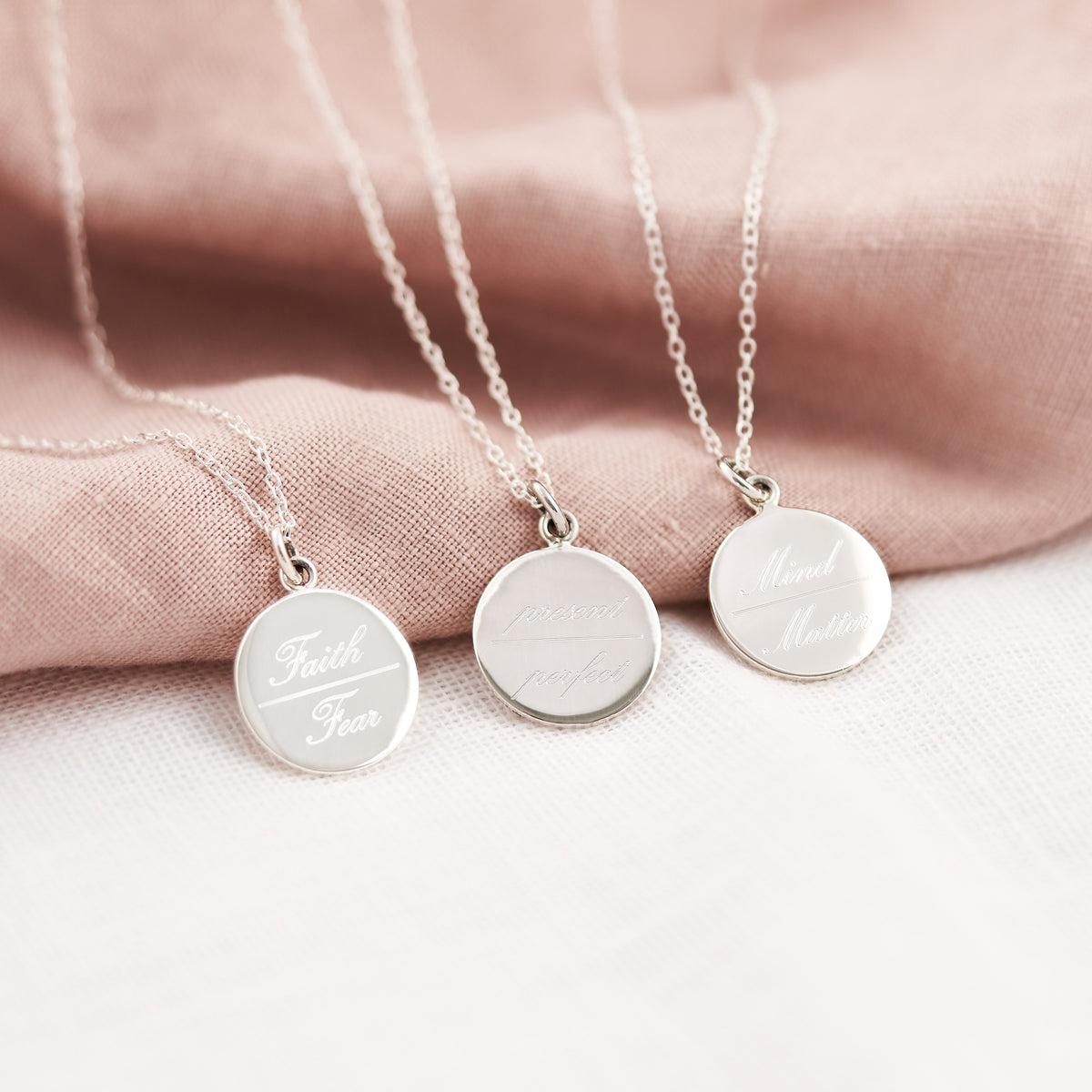 Three silver round pendant necklaces with engraved text, displayed on a soft pink fabric drape over a white surface.