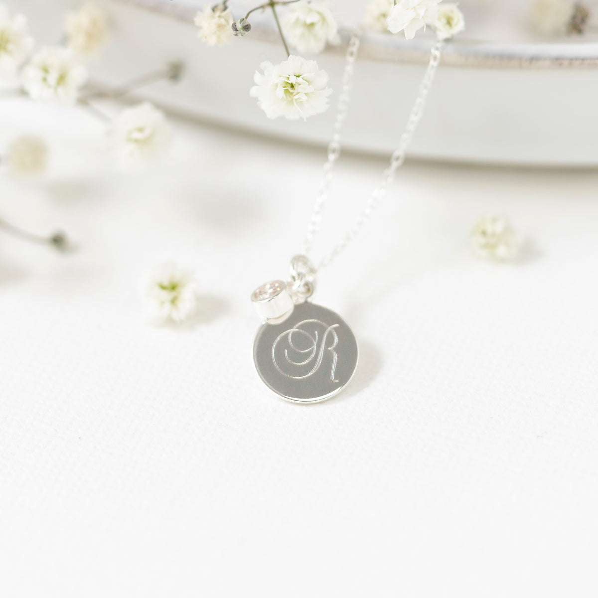 A silver initial pendant necklace with a monogram letter 'R' engraved on a round disc, hanging from a delicate silver chain. The necklace is styled with small white baby's breath flowers on a clean white background.