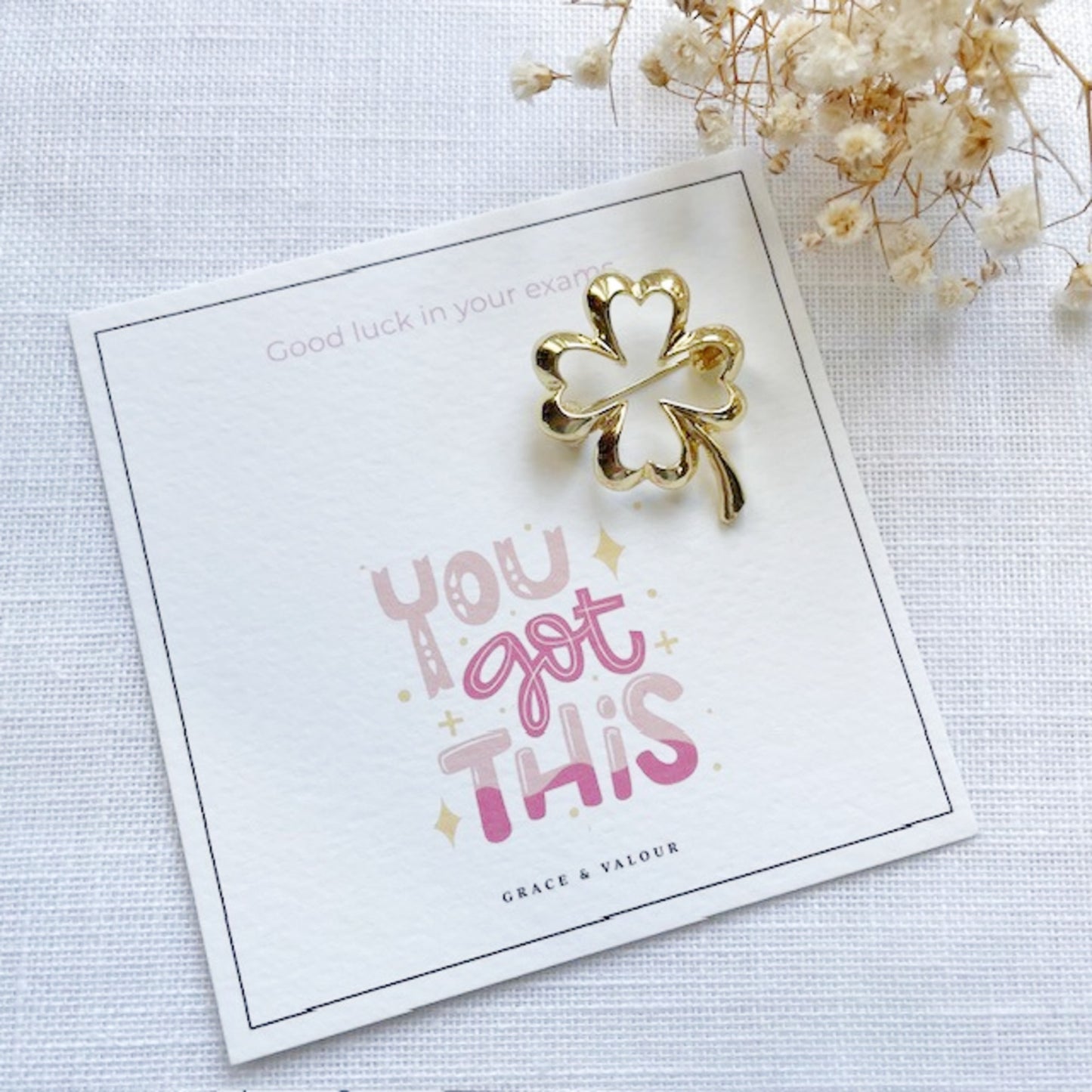 Gold four-leaf clover brooch on a card with 'You got this' text, surrounded by dried flowers.