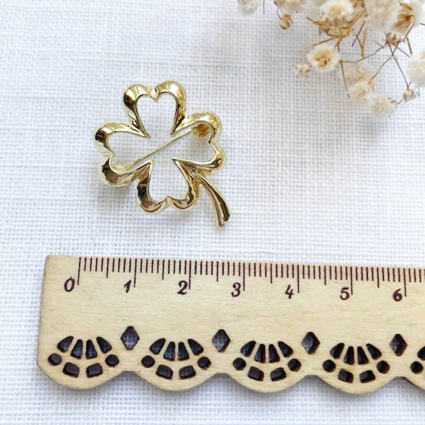 Gold four-leaf clover brooch next to a wooden ruler on a white background with dried flowers.