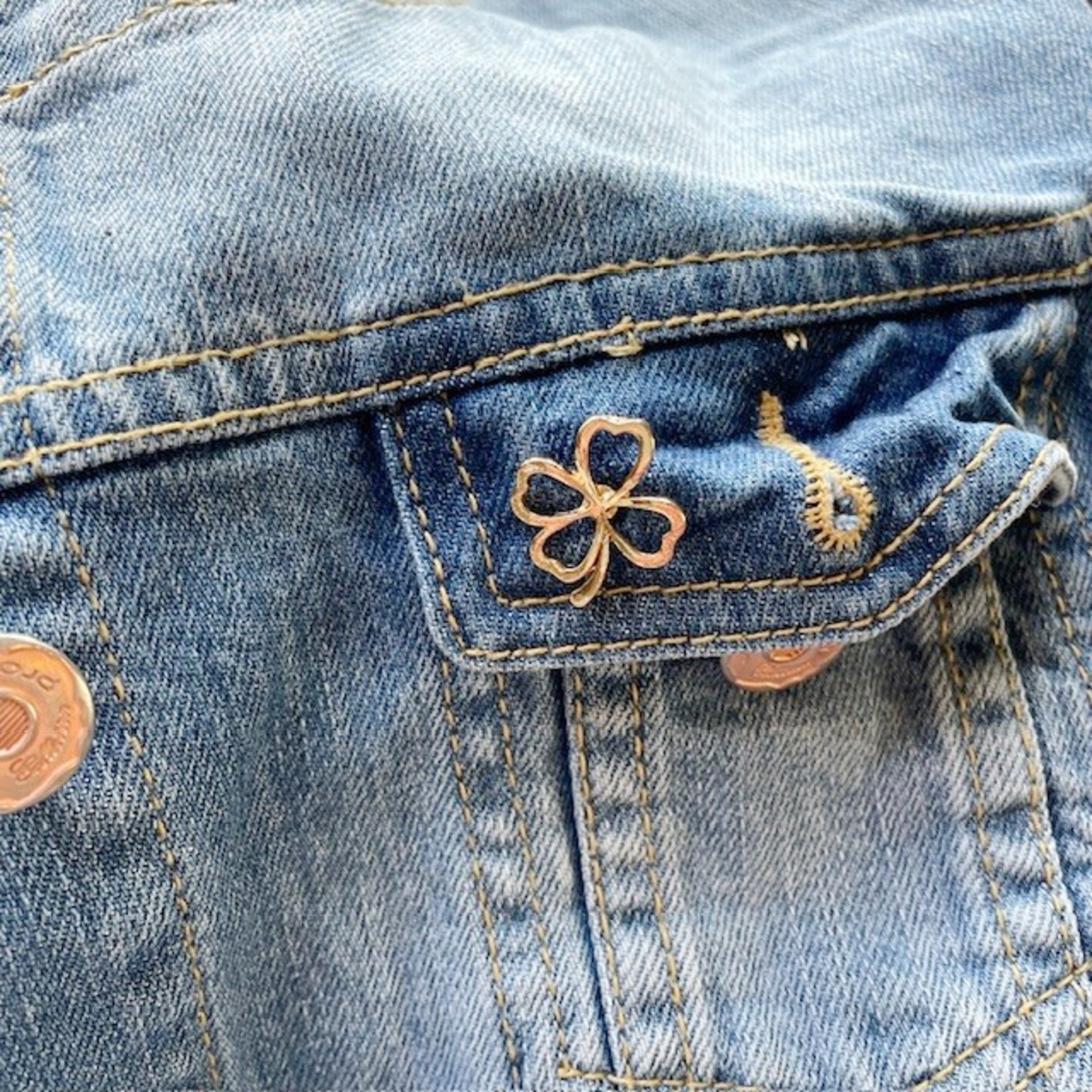 Close-up of a blue denim jacket with a gold lucky clover pin on the pocket.