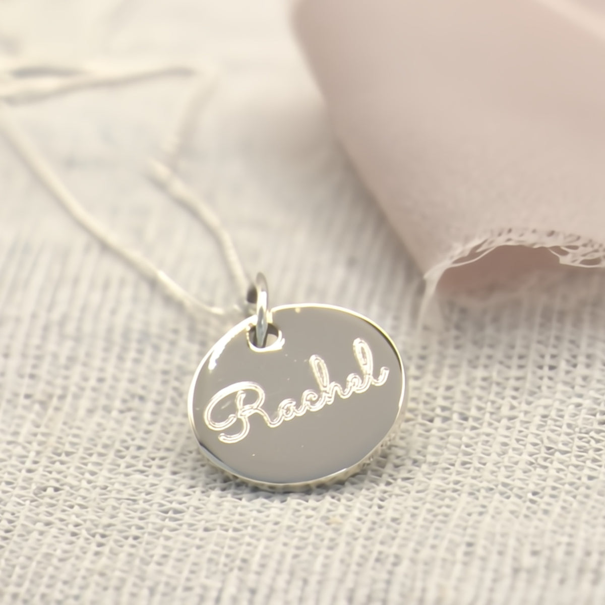 A silver oval pendant necklace engraved with the name 'Rachel' in cursive, resting on a textured white linen fabric next to a soft pink ribbon.